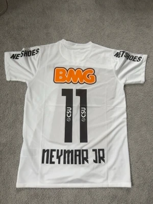 Vit Santos Neymar Jr matchtröja - Säljer en vit Santos matchtröja med Neymar Jr och nummer 11 på ryggen. Tröjan har tryckta sponsorer som BMG, Netshoes och Seara samt Santos klubbmärke på bröstet. Tillverkad i lätt och ventilerande material, perfekt för fotboll eller träning. Nyskick