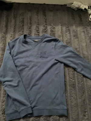 Blå sweatshirt med New York-brodyr - Mysig blå sweatshirt med broderad text 'New York' på bröstet. Tröjan har rund halsringning, långa ärmar och ribbade muddar vid ärmslut och nederkant. Perfekt för en avslappnad och stilren look.