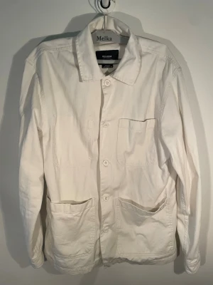 Vit overshirt från pull&bear - Stilren vit overshirt från pull&bear  med klassisk krage, tre framfickor och knäppning framtill. Tillverkad i ett mjukt bomullstyg som ger en avslappnad look. Perfekt att bära över en t-shirt eller hoodie för en enkel och clean stil.