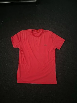 Röd kortärmad t-shirt från BOSS - Snygg röd t-shirt från BOSS med rund halsringning och diskret logga på bröstet. Klassisk passform och korta ärmar, perfekt till jeans eller shorts. Tillverkad i mjuk bomull för skön känsla hela dagen.