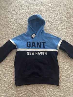 Blå hoodie från GANT med tryck - Snygg hoodie från GANT i två nyanser av blått med vita detaljer. Stor GANT-logga och texten 'NEW HAVEN' framtill. Klassisk huva och stor magficka. Perfekt för dig som gillar sportig och avslappnad stil. Helt ny med prislapp, storlek 176 vilket motsvarar S.