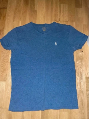 Polo Ralph Lauren T-shirt med V-ringning för män 👕 - En klassisk T-shirt med V-ringning från Polo Ralph Lauren för herrar i en mångsidig, melerad blå nyans, prydd med den ikoniska broderade logotypen (polospelaren) på vänster bröst. Tröjan är tillverkad i en skön bomullsblandning och är i utmärkt begagnat skick utan synliga defekter. ⭐️ Skick: 8/10 🎨 Färg: Melerad blå 📦 Snabb och omsorgsfull frakt