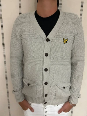Ljusgrå kofta från Lyle & Scott - Snygg ljusgrå stickad kofta från Lyle & Scott med deras klassiska gula logga på bröstet. Koftan har v-ringning, fem knappar framtill och två fickor med knappdetaljer. Ribbstickade muddar och nederkant ger en schysst passform. Perfekt till jeans eller chinos. Cardigan