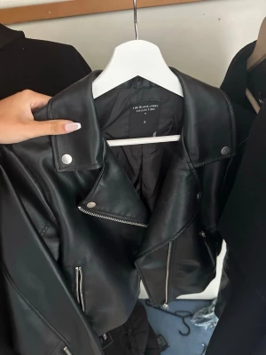 Svart skinnjacka med dragkedjor - Snygg svart skinnjacka från The Black Label Collection i storlek S. Klassisk bikerstil med silvriga dragkedjor, axelklaffar och detaljerade sömmar. Perfekt för en cool och trendig look.