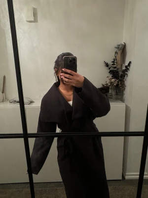 Svart kappa med knytskärp - Snygg svart kappa med bred krage och knytskärp i midjan. Kappan har en rak passform och långa ärmar, vilket ger en stilren och modern look. Perfekt för dig som vill ha en enkel men trendig ytterjacka till hösten eller vintern.