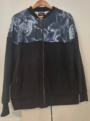 Svart hoodie med blommigt mönster - Snygg svart hoodie med dragkedja och ett unikt blommigt mönster i gråskala över axlar och bröst. Tröjan har ribbade muddar, två fickor framtill och en klassisk rak passform. Perfekt för dig som vill sticka ut med en cool detalj på din outfit.