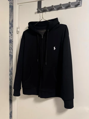 Polo Ralph Lauren zip hoodie - Klassisk svart hoodie från Polo Ralph Lauren med dragkedja framtill, huva med snörning och två fickor. Ikoniska vita logon broderad på bröstet. Perfekt för en chill och stilren look.