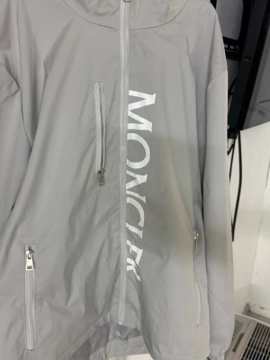Ljusgrå vindjacka från Moncler - Snygg ljusgrå vindjacka från Moncler med dragkedja framtill, två sidofickor med dragkedja och en bröstficka. Stor Moncler-logga tryckt vertikalt på framsidan och klassisk patch på ärmen. Jackan har huva och är tillverkad i ett lätt, vindtätt material. Kan gå ner i pris vid snabb affär.