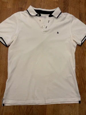 Vit pikétröja från Jack & Jones - Klassisk vit pikétröja från Jack & Jones med svart krage och svarta detaljer på ärmsluten. Tröjan har två knappar vid halsen och en liten broderad logga på bröstet. Perfekt till jeans eller shorts för en clean look.