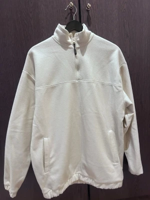 Vit/beige fleece-tröja med half zip - Mysig vit fleece-tröja med hög krage och half zip framtill. Tröjan har två sidofickor och resår i nederkant för en skön passform. Perfekt för kyliga dagar och enkel att matcha med det mesta.