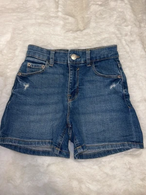 Blå jeansshorts med hög midja - Snygga blå jeansshorts med hög midja och klassisk femficksmodell. Shortsen har slitna detaljer framtill, bälteshällor och knappstängning. Tillverkade i denim med raka ben och normal längd.