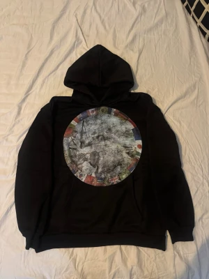 Svart hoodie Peaceinwar - Säljer en svart hoodie med stor rund grafisk print på ryggen. Trycket har ett collage av flaggor runt kanten och ett svartvitt motiv i mitten. Hoodien har klassisk huva och ribbade muddar. Perfekt för dig som gillar streetwear och unika prints.