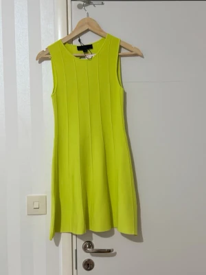 Ärmlös limegrön klänning - Snygg ärmlös klänning i en stark limegrön färg. Klänningen har en rund halsringning och vertikala sömmar som ger en smickrande form. Baksidan har en stor oval cut-out för en cool detalj. Materialet ser stretchigt och tjockare ut, perfekt för trendiga looks.