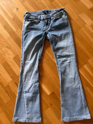 Ljusblå bootcut jeans - Säljer ett par ljusblå bootcut jeans med klassisk femficksdesign och snygga detaljer på bakfickorna. Jeansen har utsvängda ben och är tillverkade i mjukt denimtyg som ger en avslappnad look. Perfekta för dig som gillar retrovibbar och vill ha ett par jeans som sticker ut.