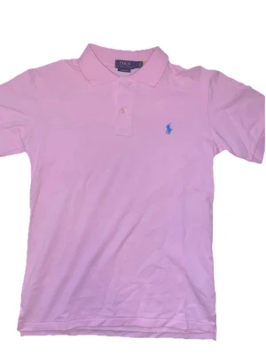 Ljusrosa piké från Polo Ralph Lauren - Klassisk ljusrosa pikétröja från Polo Ralph Lauren med korta ärmar och blå broderad logga på bröstet. Tröjan har krage och två knappar framtill. Perfekt till en avslappnad men ändå stilren look.