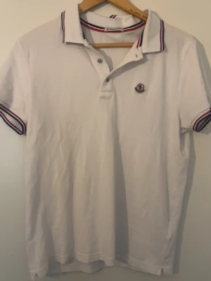 Moncler pike - Vit pikétröja med korta ärmar och krage. Ärmsluten har rödblå randiga detaljer och det finns en liten broderad logotyp på bröstet. Tröjan har knappar vid halsen och är gjord i mjukt bomullstyg. Den är storlek M sitter ungefär som S.