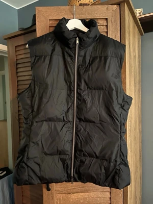 Svart dunväst med hög krage - Stilren svart dunväst med hög krage och dragkedja framtill. Västen har en quiltad design och två sidofickor med dragkedja. Perfekt för lager-på-lager och ger en cool look till din outfit.