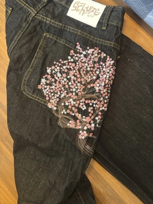 Derschutze - Jeans - Unika mörkblå jeans med snyggt broderat körsbärsträd i rosa och vitt på bakfickan samt gren med blommor på framfickan. Klassisk femficksmodell med kontrastsömmar. Perfekta för dig som vill sticka ut med coola detaljer. Har ingen hål på midjan, den får du göra själv så du kan knäppa annars har den inga defekter
