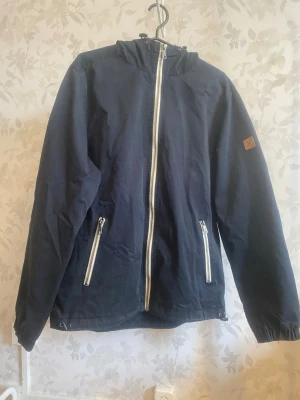 Lyle & Scott med huva och dragkedja framtill.  - Lyle & Scott med huva och dragkedja framtill. Snygg mörkblå vindjacka med huva och vita dragkedjor framtill och på fickorna. Jackan har en enkel design med resår vid ärmsluten och en liten patch på ena ärmen. Perfekt för blåsiga dagar och enkel att matcha till olika outfits.