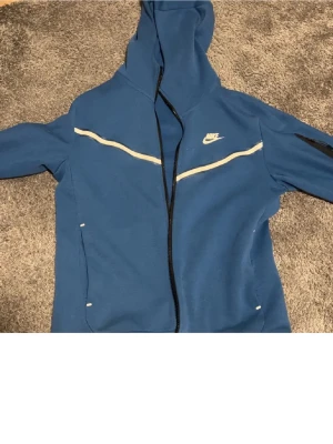 Blå Nike tech fleece  - Snygg blå hoodie från Nike med hel dragkedja framtill och vit logga på bröstet. Hoodien har vita detaljer över bröstet och en huva. Perfekt för en sportig och avslappnad stil.