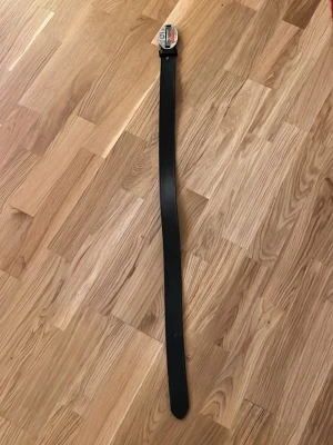 Svart klassiskt Harley Davidson bälte i skinn, 92cm - Snyggt och stilrent svart bälte i skinn med metallspänne. Perfekt för att lyfta vilken outfit som helst och funkar till både jeans och kostymbyxor. Enkel design som passar till det mesta.