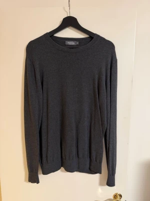 Stickad tröja Dressmann - En enkel och stilren mörkgrå stickad tröja med rund halsringning och långa ärmar. Tröjan har ribbade muddar vid ärmslut och nederkant. Perfekt basplagg till garderoben och lätt att matcha med olika outfits.