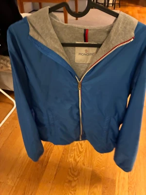 Blå vindjacka från Moncler med huva - Snygg blå vindjacka från Moncler med grå huva och dragkedja framtill. Jackan har lång ärm och klassisk Moncler-logga på ärmen. Perfekt för dig som vill ha en stilren och sportig look. Tillverkad i lätt polyester.