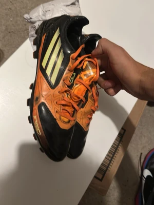 Adidas F10 TRX shoes 2011 - Säljer ett par snygga fotbollsskor från Adidas i svart och orange med gula detaljer och klassiska tre ränder på sidan. Skorna har fasta dobbar och snörning framtill. Perfekta för dig som vill sticka ut på planen!