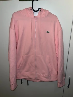 Rosa zip hoodie från Lacoste - Snygg rosa hoodie från Lacoste med dragkedja framtill, huva med dragsko och klassisk krokodil-logga på bröstet. Tillverkad i mjukt bomullsmaterial och har två stora fickor framtill. Perfekt för en chill och avslappnad stil. Passar både M och L
