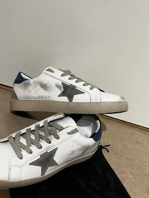Vita sneakers med grå stjärna - Snygga vita sneakers med grå stjärndetalj på sidan, grå skosnören och blå häl. Skorna har en klassisk låg profil och är tillverkade i läder med slitna detaljer för en trendig look.