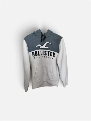 Gråvit hoodie från Hollister - Snygg hoodie från Hollister i grått och vitt med stor logga och text på bröstet. Hoodien har huva med dragsko och en stor magficka. Tillverkad i mjukt material, perfekt för chill dagar. Klassisk passform och stilren design.