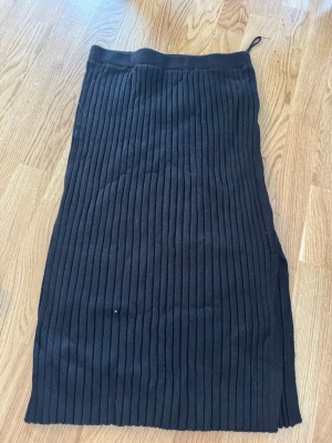 Svart ribbad långkjol med slits - Säljer en svart långkjol med ribbad struktur och slits på sidan. Kjolen har en enkel och stilren design som passar till många olika looks. Perfekt för dig som gillar en clean och modern stil.
