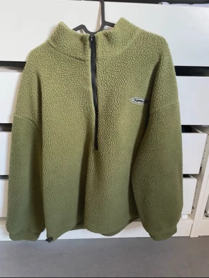 Olivgrön fleece-tröja med halv dragkedja - Mysig olivgrön fleece-tröja med hög krage och halv dragkedja framtill. Tröjan har en relaxed passform, långa ärmar och ett litet broderat märke på bröstet. Perfekt för lager-på-lager och chill dagar.