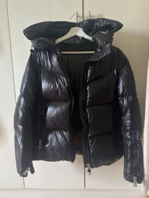 Svart Vinterjacka - Snygg svart dunjacka med glansig finish och stor huva med vit logga. Jackan har dragkedja framtill, två fickor med dragkedja och ribbade muddar vid ärmsluten. Perfekt för kalla dagar och riktigt trendig look.