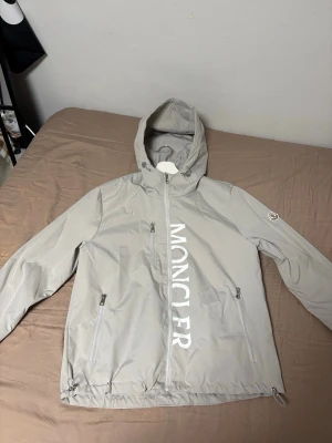 Ljusgrå vindjacka från Moncler - Snygg ljusgrå vindjacka från Moncler med huva och dragkedja framtill. Jackan har stora vita Moncler-bokstäver på framsidan, två fickor med dragkedja och justerbar nederkant. Perfekt för blåsiga dagar och streetwear-stil.