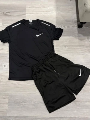 Svart träningsset från Nike - Snyggt svart träningsset från Nike med t-shirt och shorts. T-shirten har vita detaljer på axlarna och Nike-logga på bröstet. Shortsen har elastisk midja med dragsko och vit Nike-logga på benet. Perfekt för gymmet eller löpning.