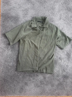 Olivgrön ribbad skjorta - Säljer en olivgrön skjorta med korta ärmar med texturerad struktur. Skjortan har klassisk krage och knappar framtill. Perfekt för dig som gillar en avslappnad men ändå stilren look.