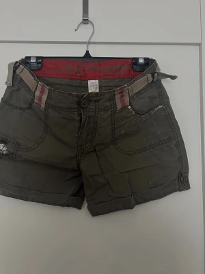 Olivgröna shorts från Ed Hardy (L) - Säljer ett par olivgröna shorts från Ed Hardy med coola detaljer och kontrastsömmar. Shortsen har fickor fram och bak, bälte i midjan och broderade detaljer. Perfekta för sommaren och ger en avslappnad streetwear-vibe.
