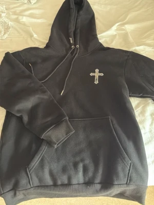Svart hoodie med korsdetalj - Säljer en svart hoodie med stor luva och magficka. På bröstet finns ett broderat kors med motiv i mitten. Hoodien passar för både män och kvinnor 