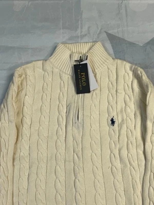 Vit kabelstickad tröja Polo Ralph Lauren - Kabelstickad vit tröja från Polo Ralph Lauren med halv dragkedja och hög krage. Tröjan har det klassiska broderade logotypen på bröstet och ribbstickade muddar. Perfekt för lager-på-lager och en stilren look.