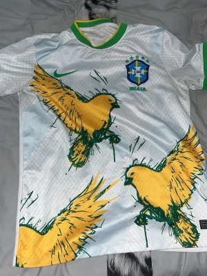 Vit Brasilien fotbollströja med fåglar - Säljer en vit fotbollströja från Nike med Brasilien-emblem på bröstet. Tröjan har stora gula fåglar tryckta över hela framsidan, gröna detaljer vid krage och ärmslut samt ett grönt Nike-märke. Perfekt för dig som älskar unik sportstil.