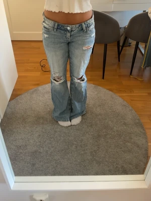 Ljusblå slitna bootcut jeans - Säljer ett par ljusblå bootcut jeans med slitningar och hål på båda knäna. Jeansen har fem fickor, normal midja och klassisk knappgylf. Perfekta för en avslappnad och trendig look.