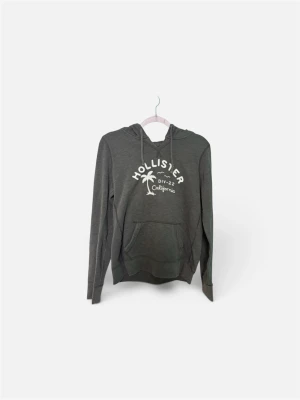 Mörkgrå Hollister hoodie med tryck - Mörkgrå hoodie från Hollister med vit brodyr framtill där det står 'Hollister DIV-22 California' och en palm. Klassisk känguruficka och dragsko i huvan. Mjuk bomullsblandning och ribbade muddar vid ärmar och nederkant.