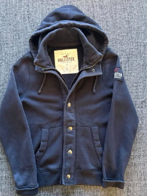  Hollister cardigan - Snygg mörkblå hoodie från Hollister med dragkedja, knappar och två stora fickor framtill. Hoodien har en stor huva med dragsko och mjukt foder. Hollister-logga på ärmen och insidan av huvan är ribbad. Perfekt för chill dagar. 