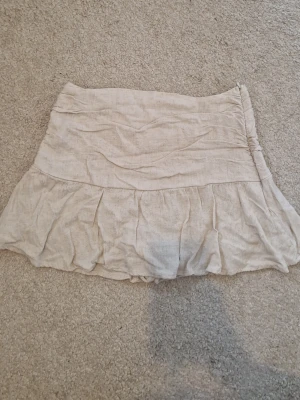 Beige kjol i linne med volang - Beige kortkjol i linne med volangkant och bred midja. Kjolen har en skort-design med shorts under för extra komfort. Perfekt för en avslappnad och trendig sommarstil. Sitter som en storlek M.