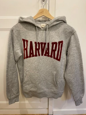Grå hoodie med HARVARD-tryck - Snygg grå hoodie med stor röd HARVARD-text i sammet på bröstet. Klassisk modell med huva, dragsnören och ficka framtill. Tillverkad i mjukt sweatshirtmaterial, perfekt för chill dagar eller plugget. 