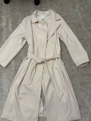Beige trenchcoat med knytskärp - Stilren beige trenchcoat med klassisk krage, knytskärp i midjan och dubbla knappar framtill. Långa ärmar och två fickor på sidorna. Perfekt för lager-på-lager och en clean look. Materialet ser ut att vara mjukt tyg, troligen bomullsblandning.