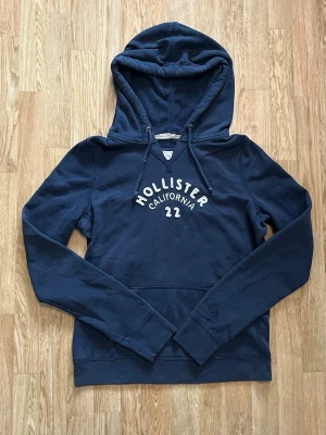 Hollister hoodie i mörkblått – Storlek L - Säljer en snygg Hollister hoodie i mörkblå/navy färg med klassiskt Hollister California 1922-tryck på framsidan. Hoodien har huva med dragsko samt en praktisk magficka.  Storlek: L Märke: Hollister Färg: Mörkblå / Navy Skick: Väldigt fint skick utan hål eller större defekter.  En stilren och bekväm hoodie som passar perfekt till vardags och går enkelt att matcha med jeans eller mjukisbyxor.  #hollister #hollisterhoodie #hoodie #sweatshirt #streetwear #navy #mörkblå #storlekl #casual #unisex