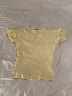 Gul ribbad topp med volangärmar - Söt ljusgul ribbad topp med volangdetaljer på ärmarna. Toppen har rund halsringning och är tillverkad i ett mjukt, stretchigt material som känns bekvämt mot huden. Perfekt för dig som vill ha en feminin och fräsch look.