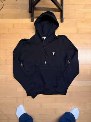 Ami Paris hoodie  - Säljer en svart hoodie med huva och dragsnören. På bröstet finns en liten vit broderad logga i form av ett hjärta med ett X. Hoodien har magficka och ribbade muddar vid ärmar och nederkant. Perfekt för en chill och stilren look.